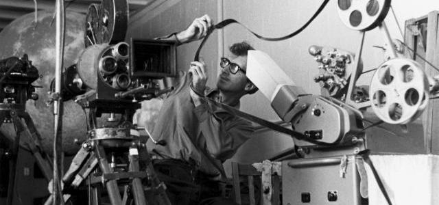 A film editor in the 1960s working on a Moviola machine, manually cutting and splicing 35mm film reels in a vintage editing room