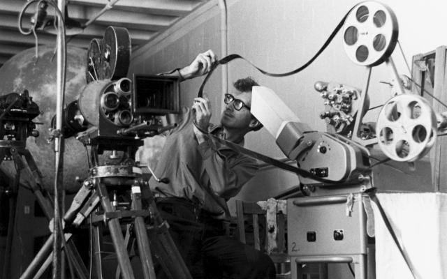A film editor in the 1960s working on a Moviola machine, manually cutting and splicing 35mm film reels in a vintage editing room